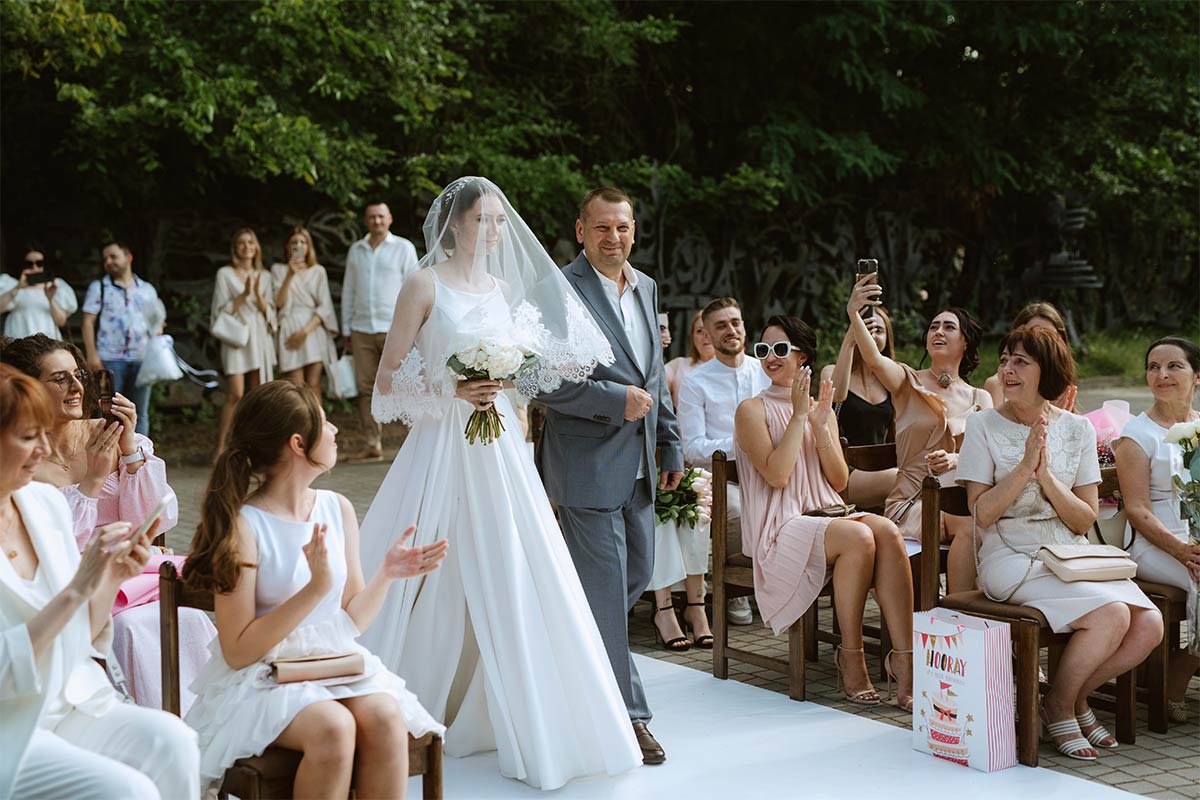 White bride with veil is walked down a white outdoor aisle by father as people clap in their seats on a sunny day