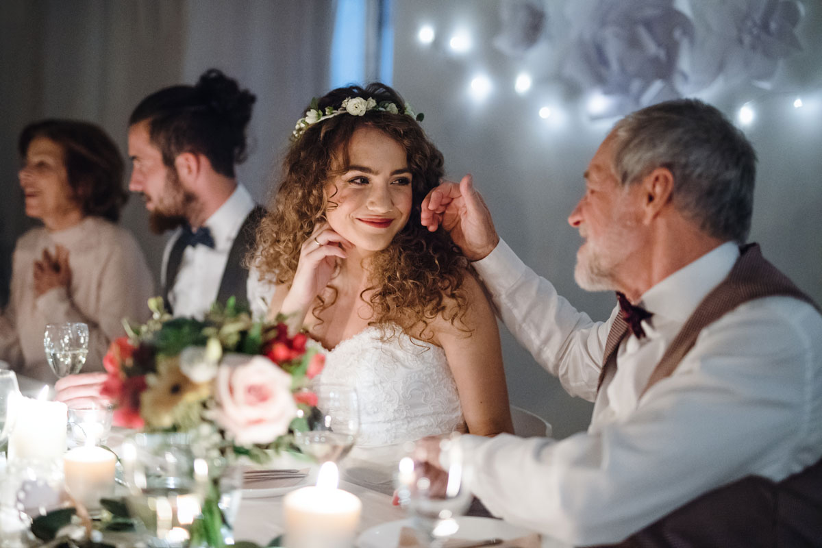 young curly-haired bride and her older father are both seated at a wedding party and looking at each other lovingly