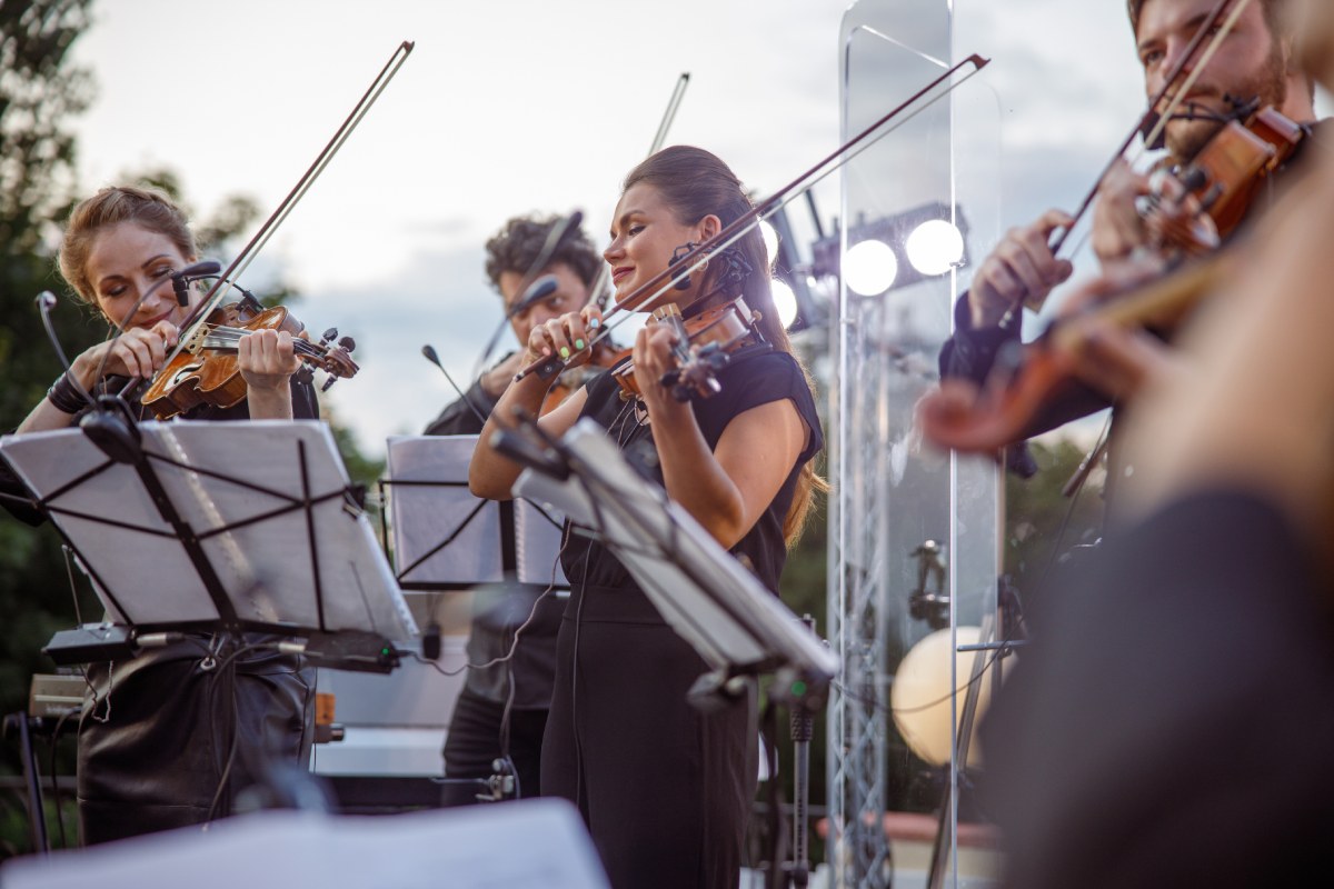 Outdoor orchestra focusing on two white women and one white male in their thirties playing violins with music stands holding sheet music