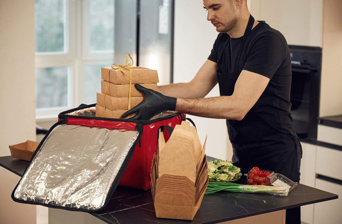 A 30 year old white man wearing a black shirt, apron, and black disposable gloves packing beige food boxes into a large red cooler, with vegetables next to the cooler on the black table top.