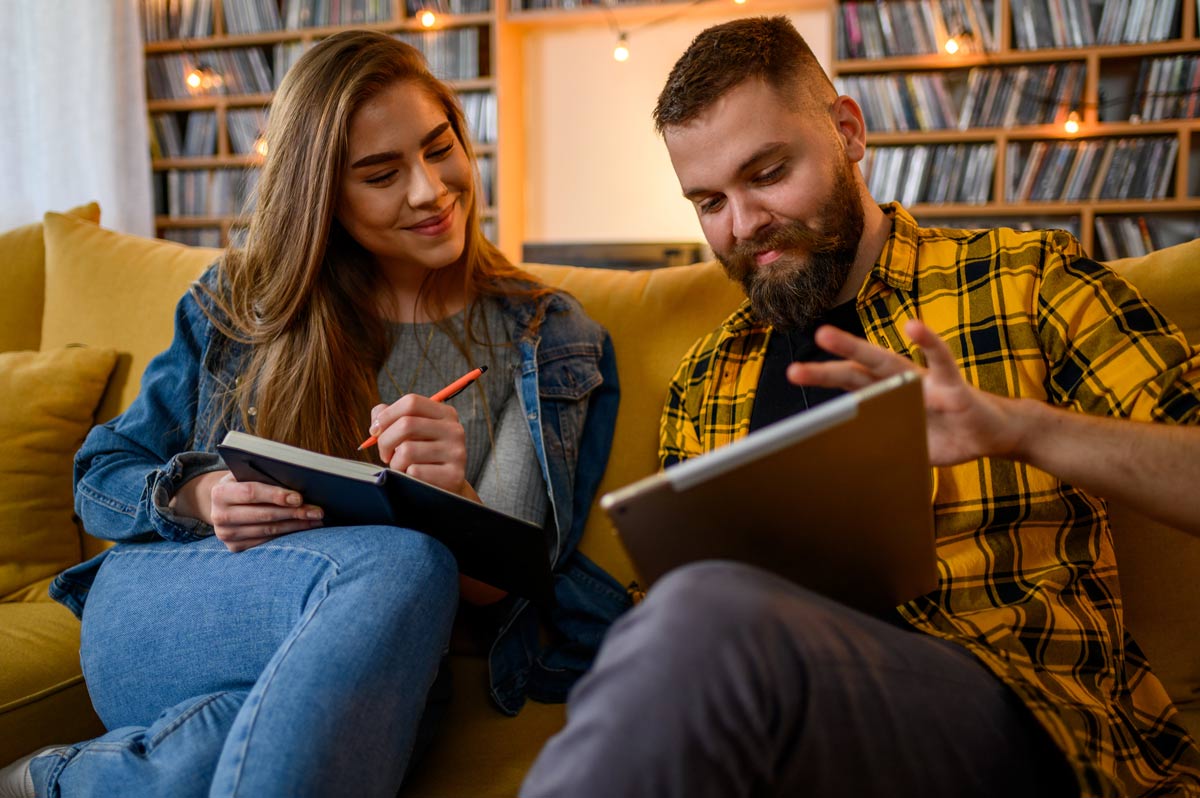 Young white couple in their late 20s using a digital tablet and a notebook while planning their wedding budget in a cozy indoor setting.