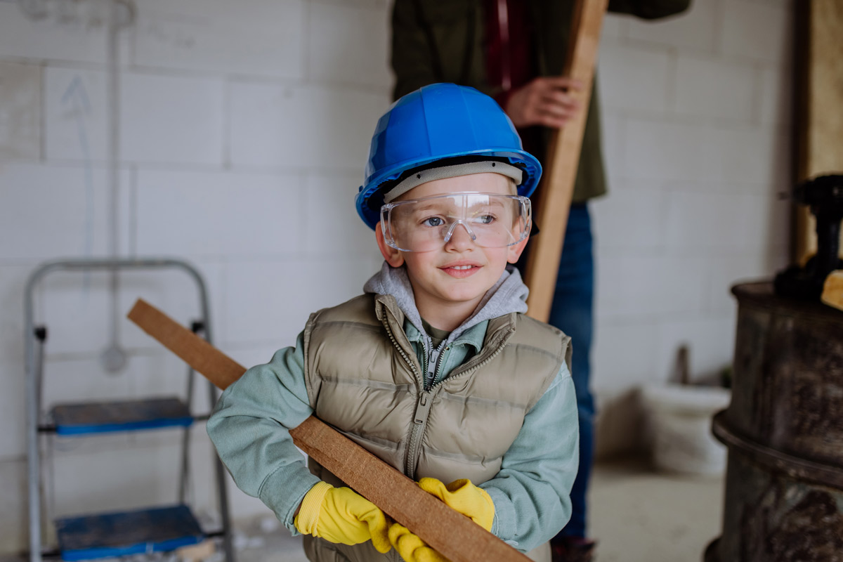 White 5 year old boy wearing construction outfit and carrying a piece of wood in white bricked building.