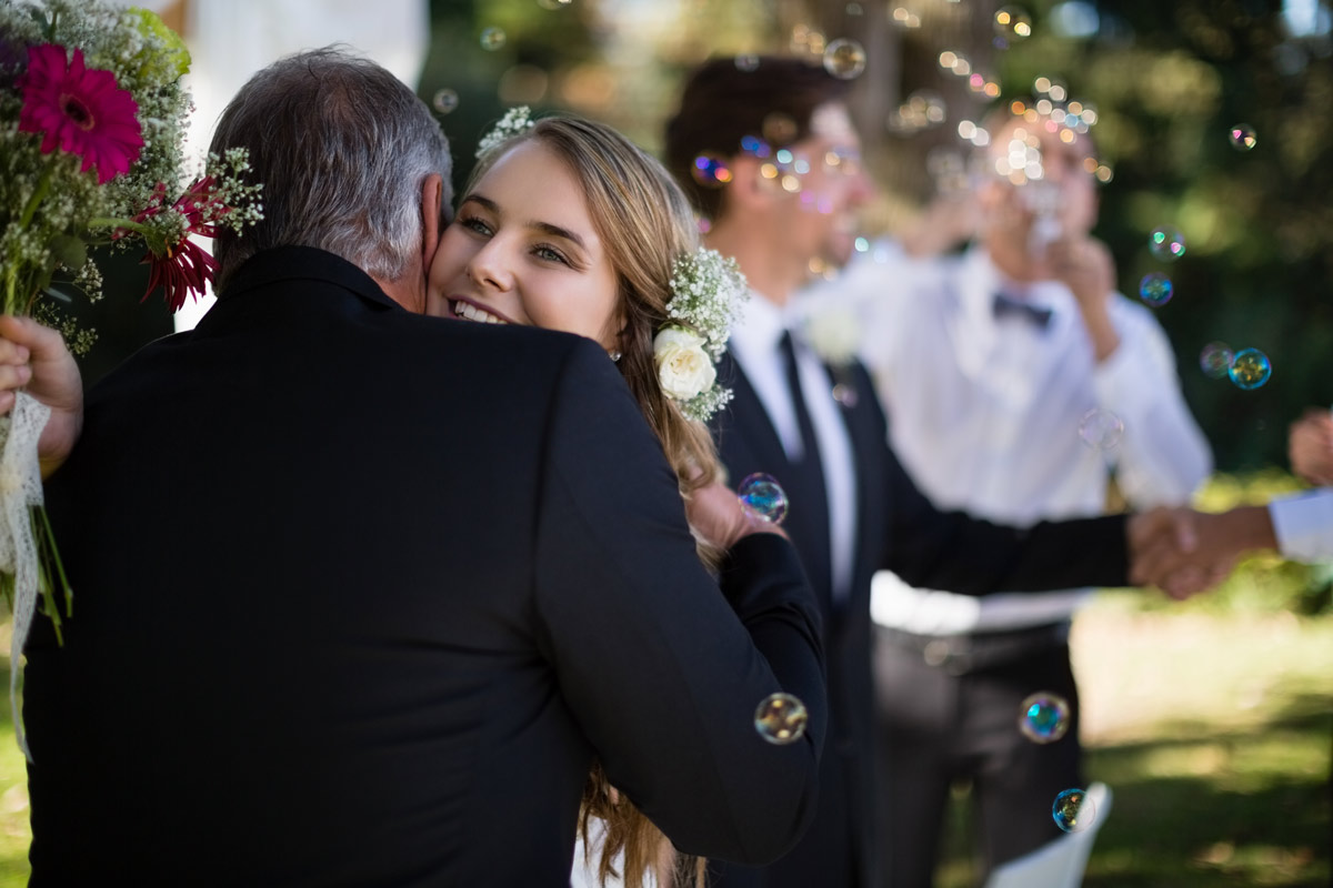 blonde bride hugging her well-dressed older father as party attendees blow bubbles