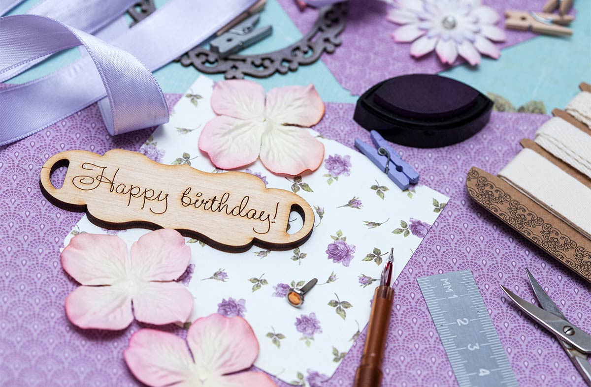 Small wooden cursive “happy birthday” sign on table surrounded by purple, pink, and blue ribbon and craft supplies 