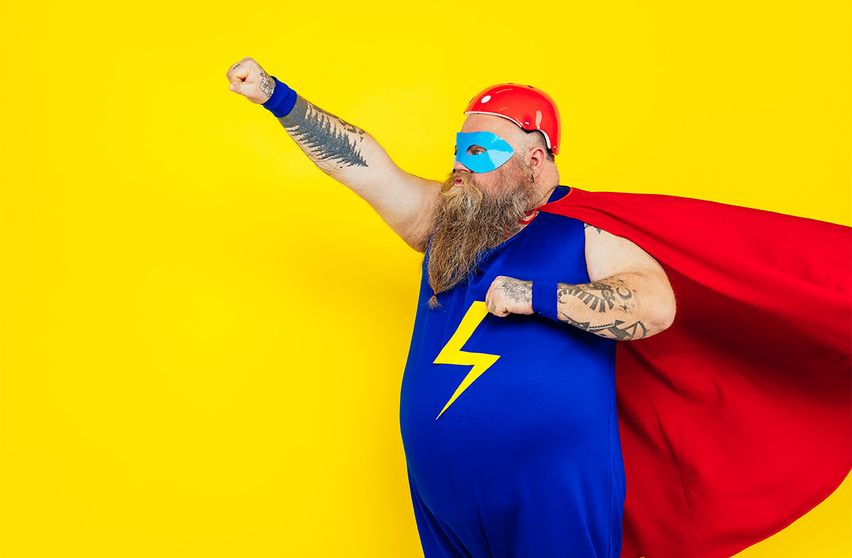 White 40-something man with long blonde beard dressed as superhero doing a superhero punch pose against a bright yellow background