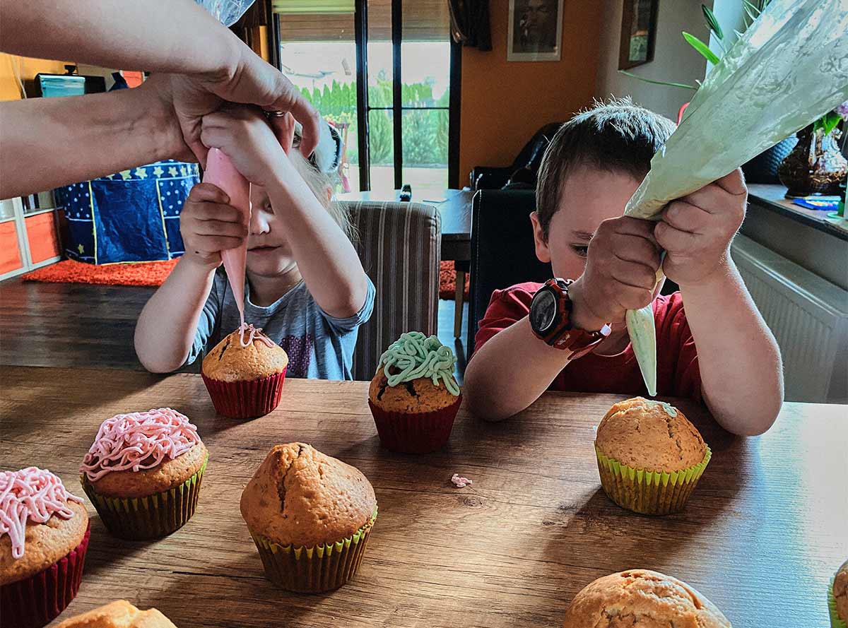 Two little white kids putting icing on cupcakes, while sitting at a wooden table in a home