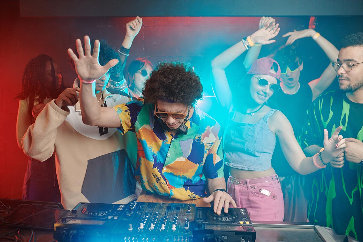 a black 25 year old  DJ wearing sunglasses dj-ing and a crowd of lively people dancing behind him