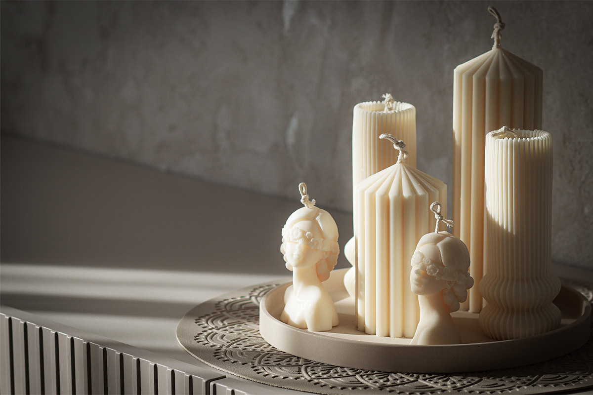 Different shaped white candles on a white tray, sitting on a gray table with a gray wall in the background. 