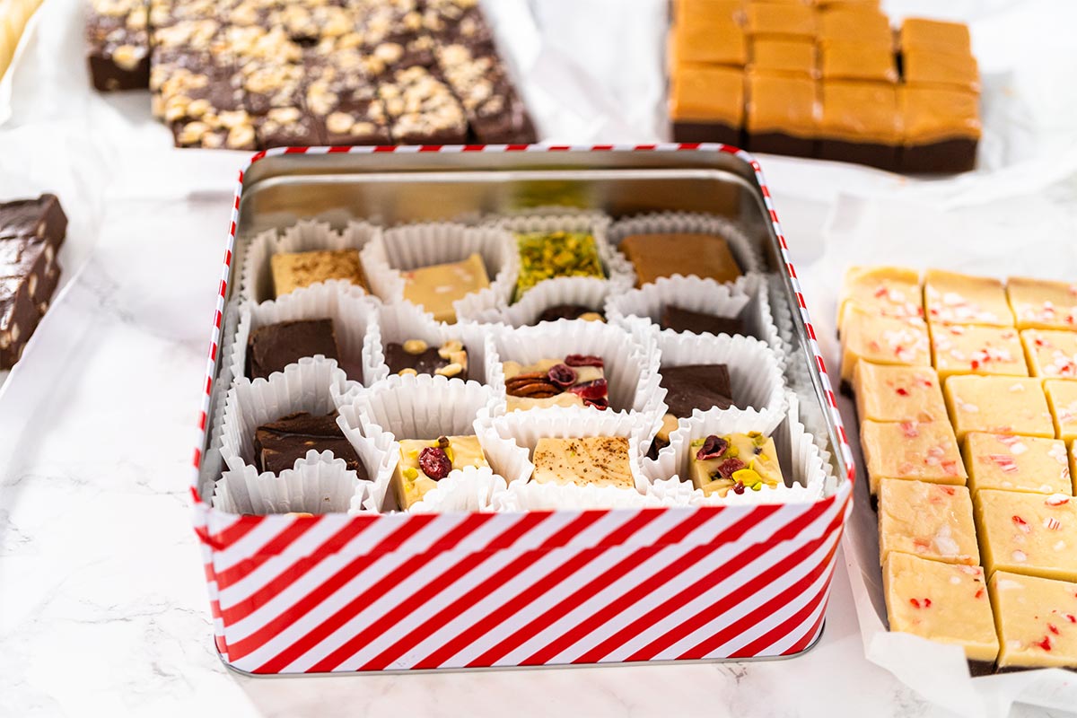 Red and white striped tin filled with different assortment of chocolates on a white table, with other chocolates in the background. 