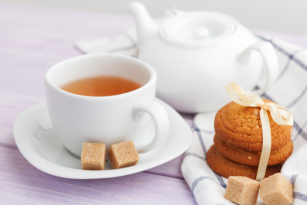 A small white mug of tea with two sugar cubes sits on a white table next to a small stack of cookies, a white tea pot, sugar cubes, and a white and navy napkin