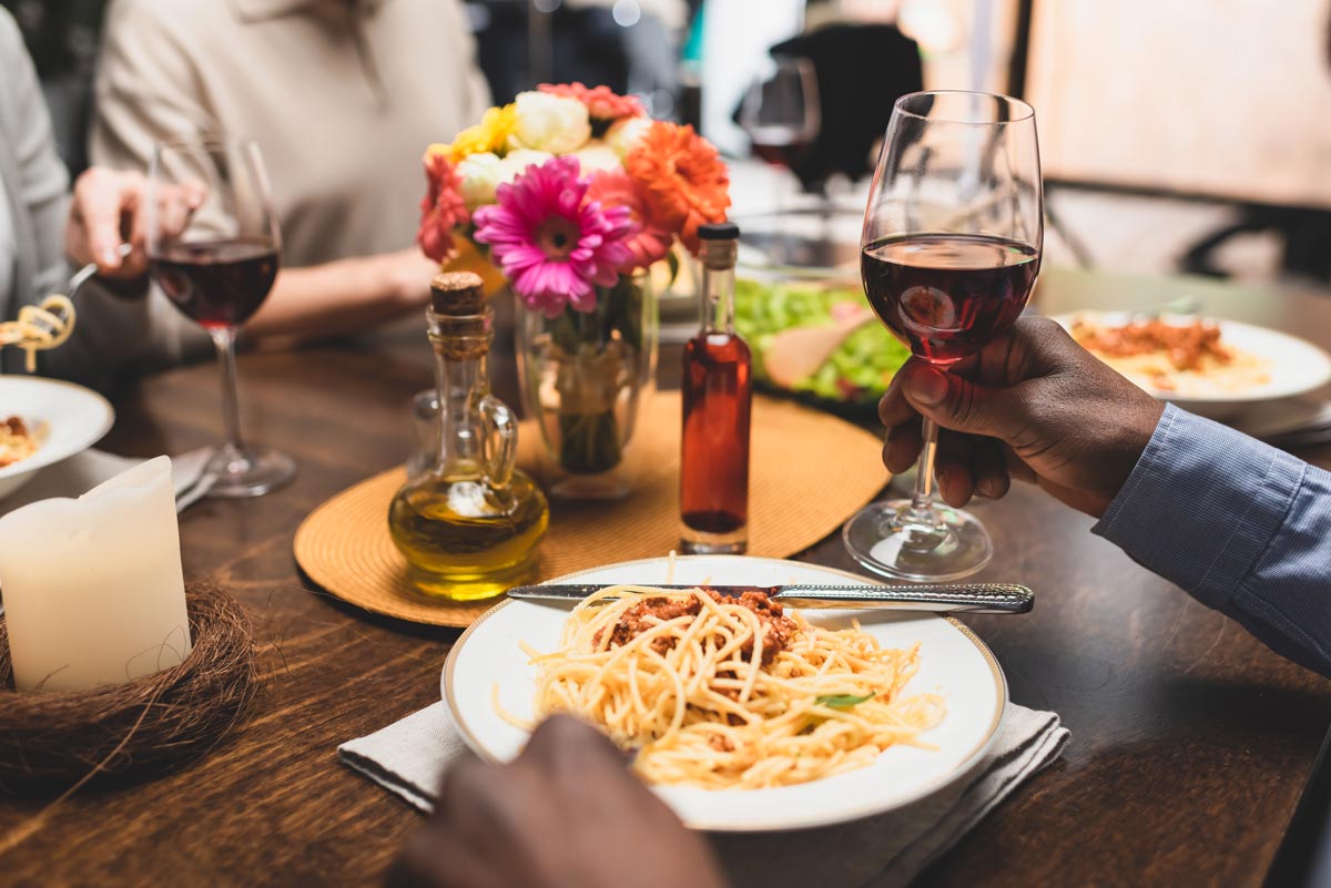 Black hand holds a glass of red wine at a table with spaghetti entree plate and centerpiece of flower vase and oil bottles 