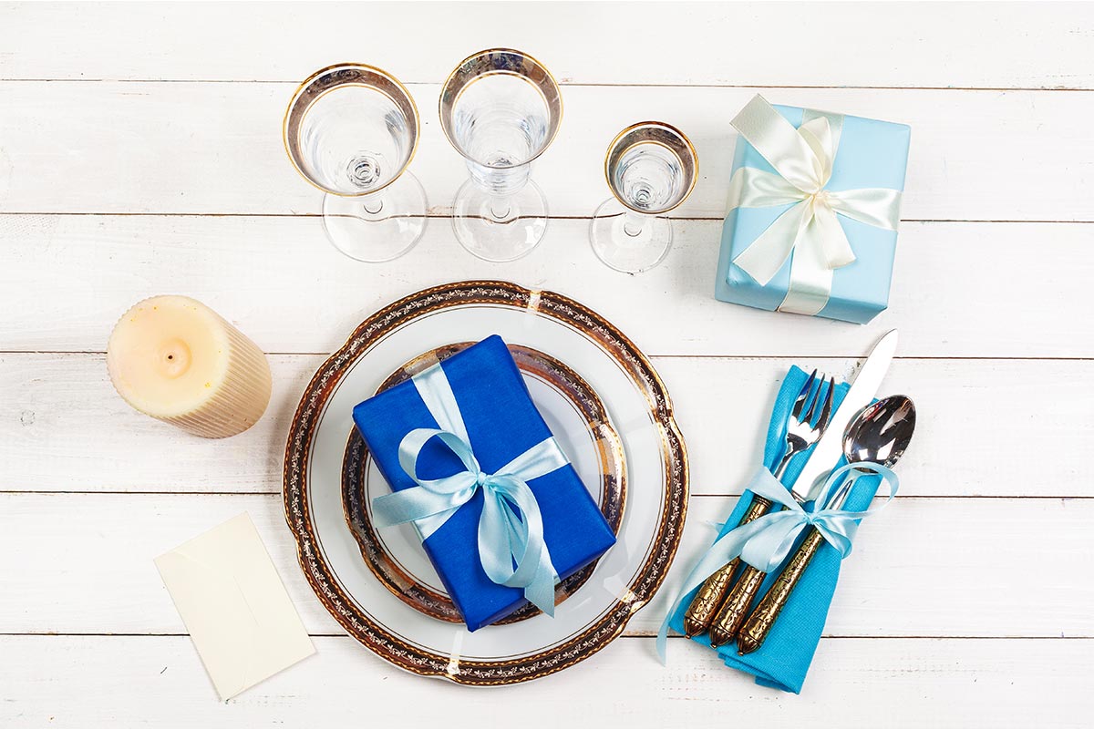 white wooden table with a luxurious gold and white plate that has a blue gift sitting on it, as well as, fancy golden utensils, a blue napkin, a light blue gift, three cups, a candle, and a card