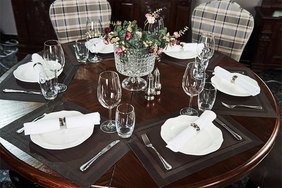 perfectly set up dark wooden table with six plaid seats contains white plates, white napkins, glasses, utensils, a fancy plant, and salt and pepper shakers