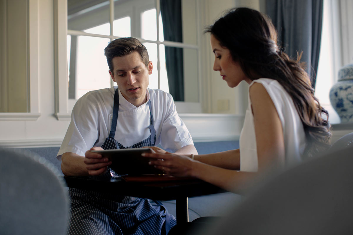 White male caterer in his early 30s reviewing menu options with a 40 year old female client on a tablet, seated at a table in a refined venue. 