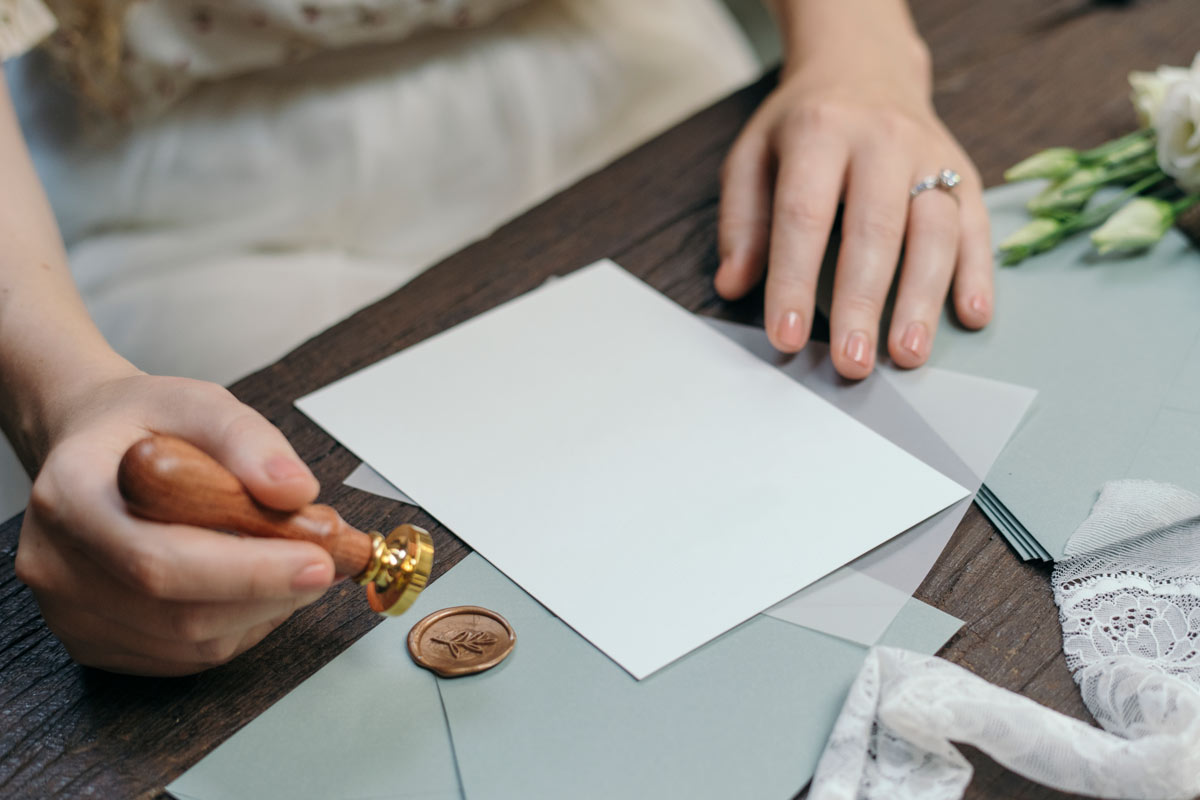 Person working on homemade wedding invitations with white paper, light blue envelopes, and a wax stamp with bronze wax.