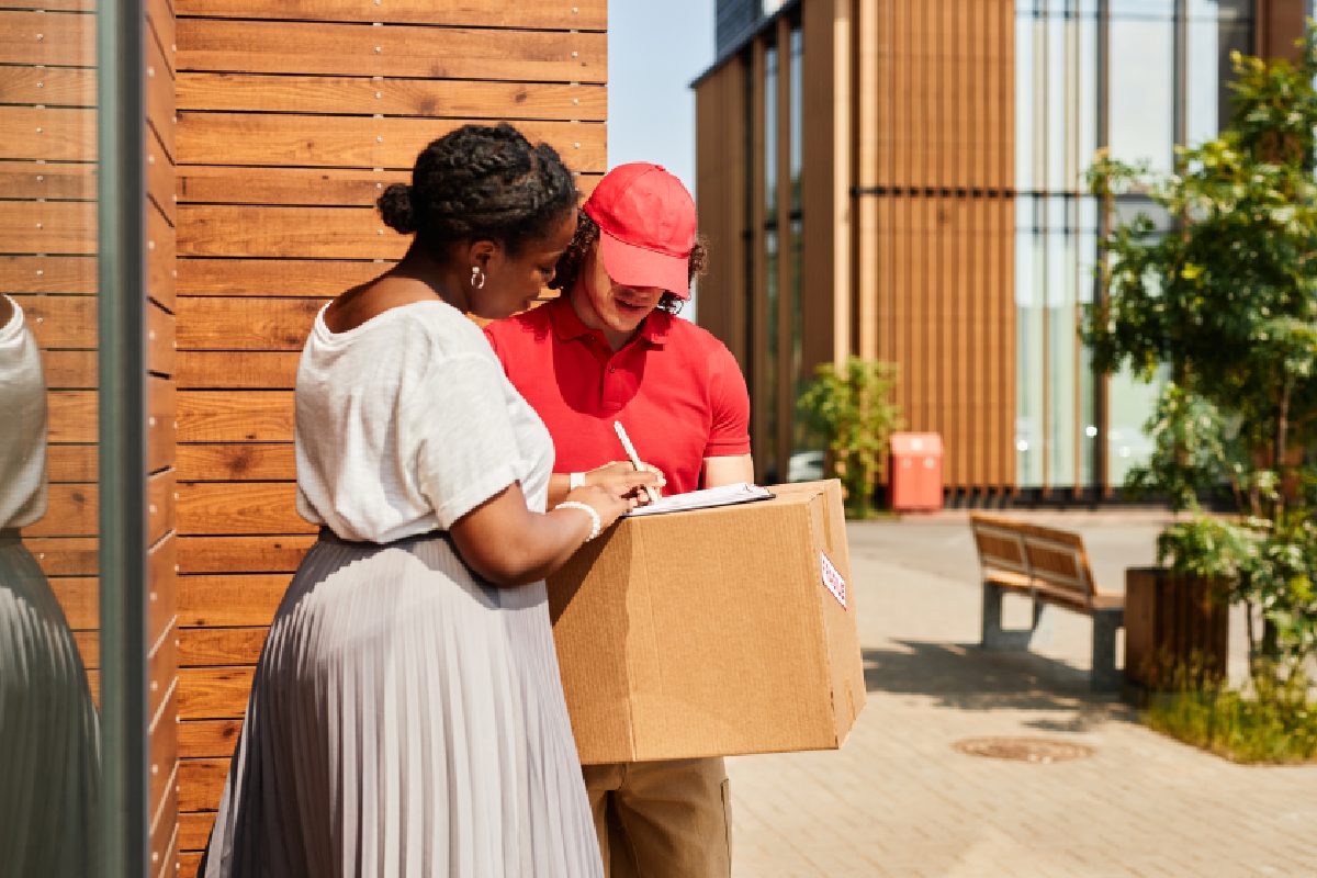 Black woman signing off on a delivery from a white man in a red hat and shirt holding a box outside of a wood paneled building.