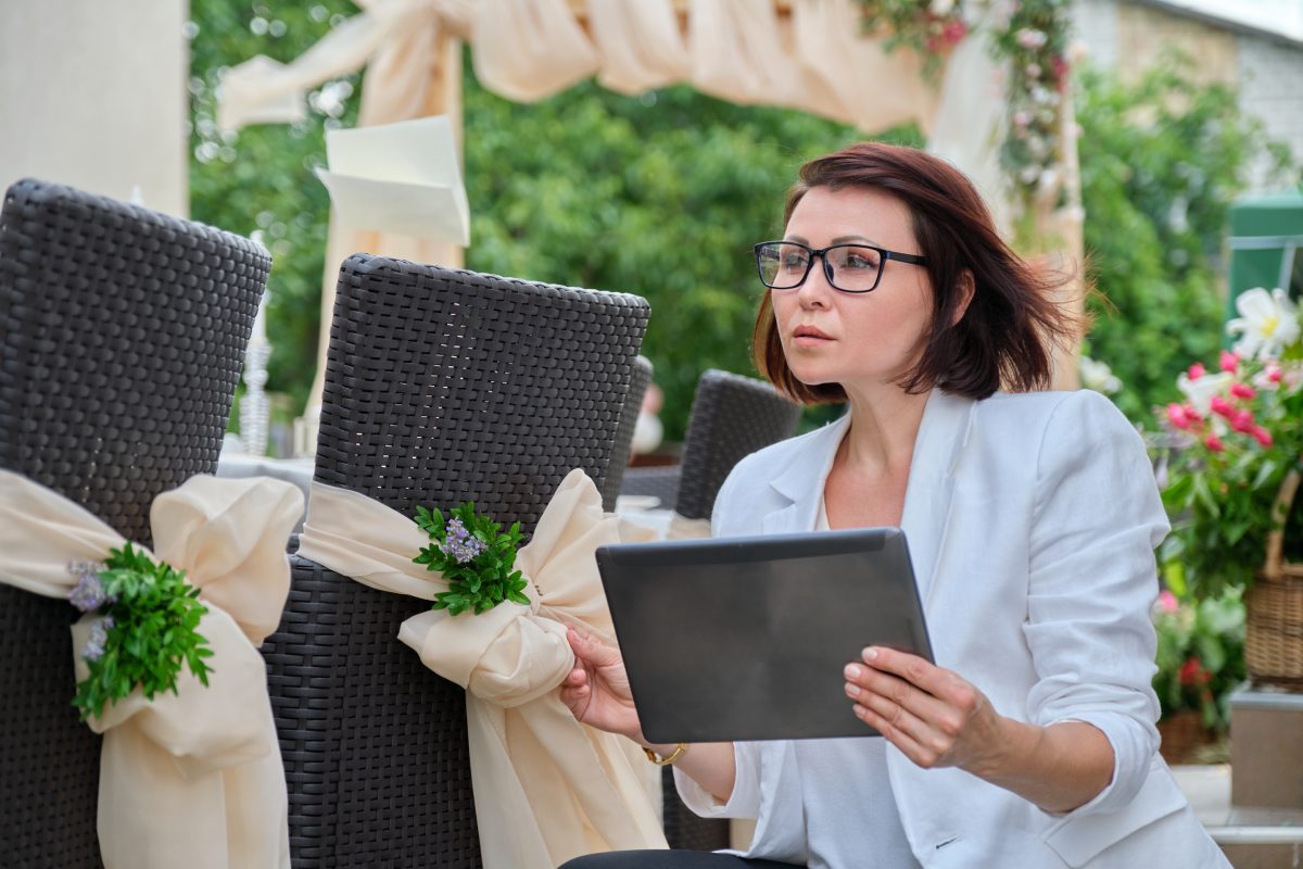 White woman wedding planner with a tablet at a wedding venue behind chairs with off-white bows and flowers.