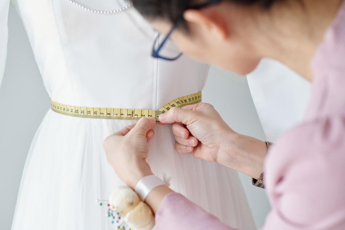 White 60-year-old woman wearing glasses altering a wedding dress on a mannequin. 