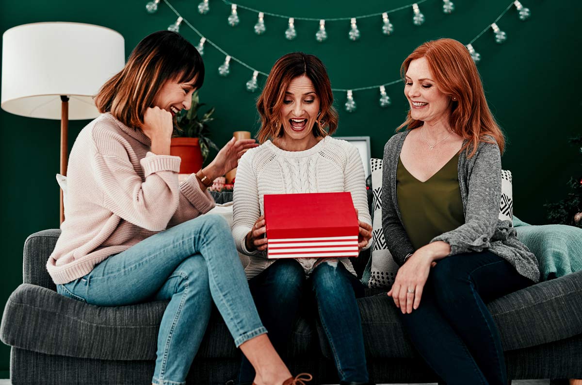 Three white 40 something women sit on a couch, the woman in the middle holds an open red gift box, looking surprised and excited, while the other two women smile at her.