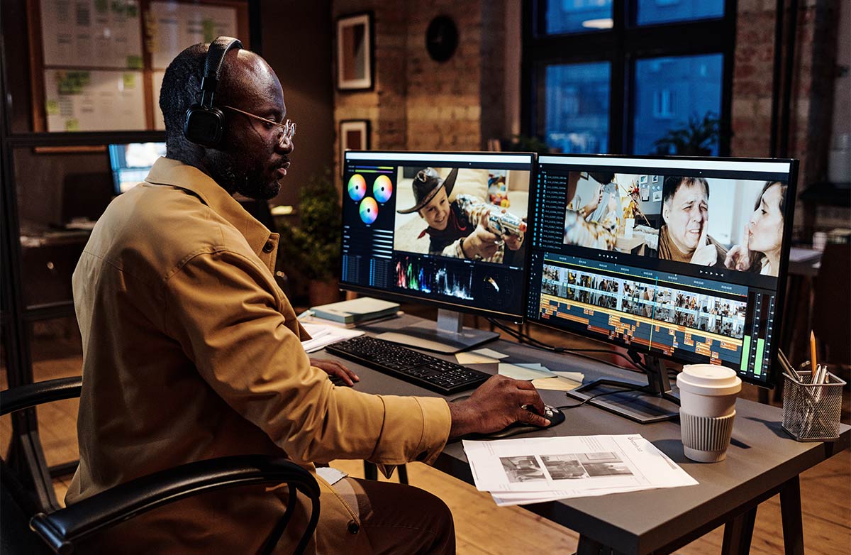 30-something black man sitting at a desk in an office in front of a desktop computer editing videos.