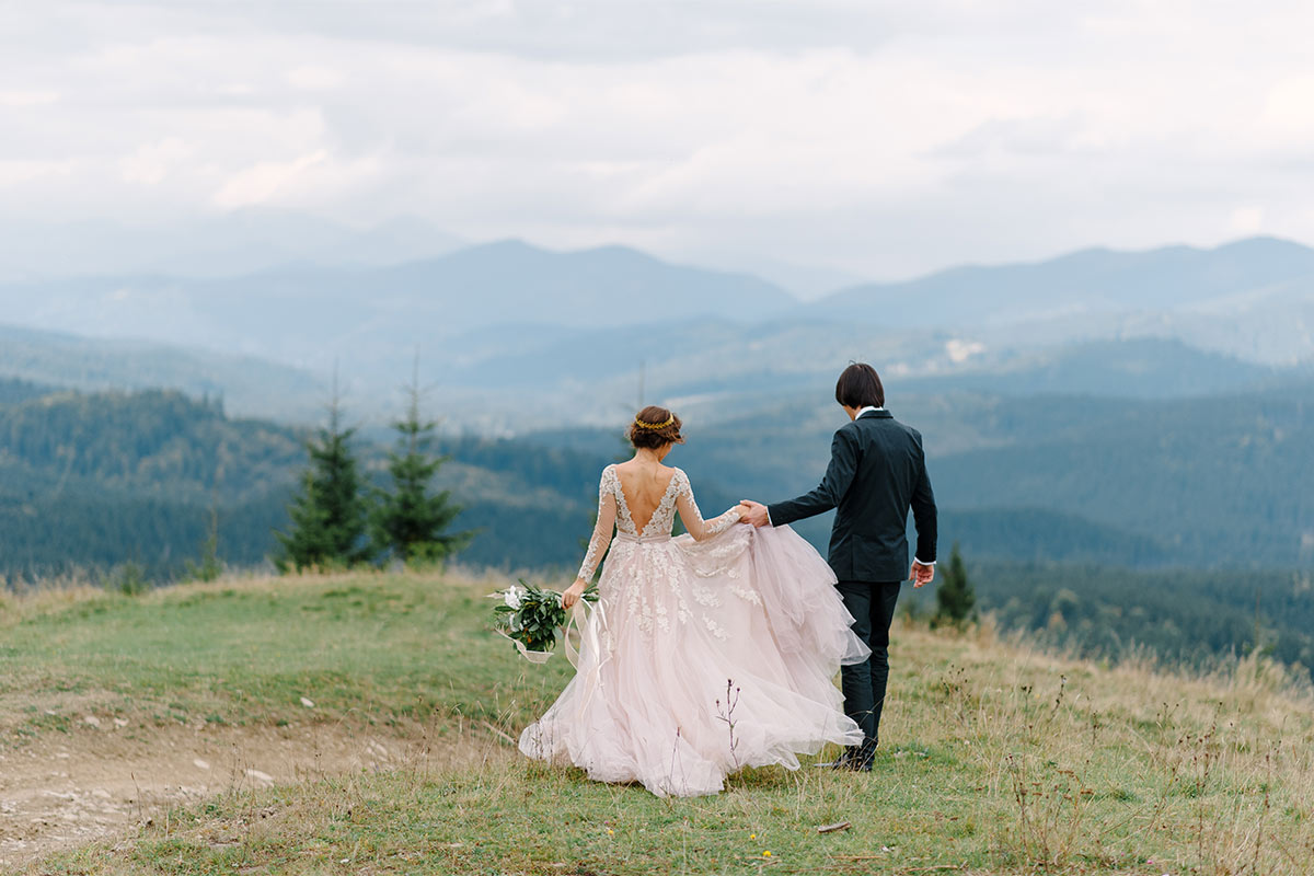 Shot from behind of a young adult white couple in wedding dress and black suit holding hands and walking with mountains and pine trees in the distance during daytime