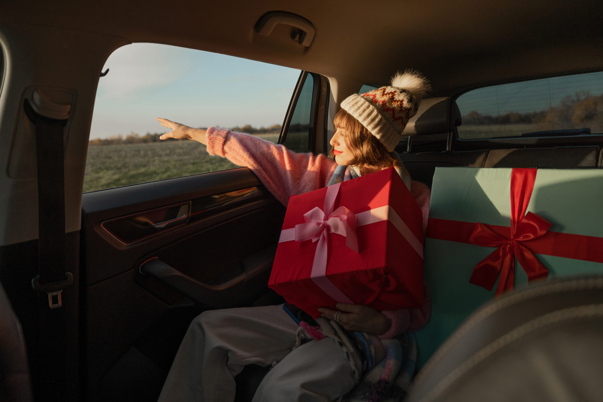 White 20 something woman wearing a stocking cap in a car surrounded by large gift-wrapped presents with bows in the backseat.