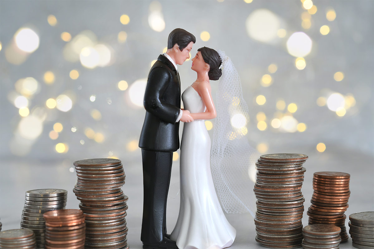  Bride and groom cake topper sits on a table surrounded by stacked coins with a blurred fairy light background 