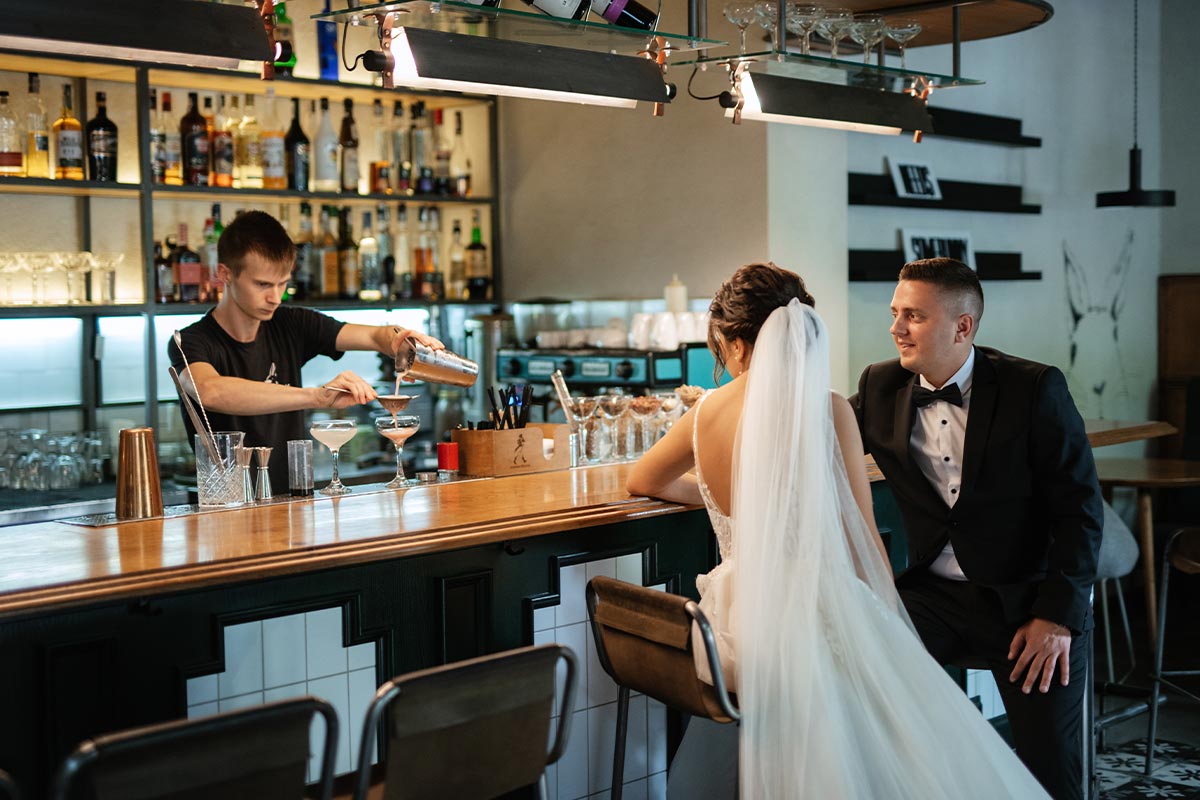 White 30 something bride and groom together inside a cocktail bar being served by a 20 something white bartender