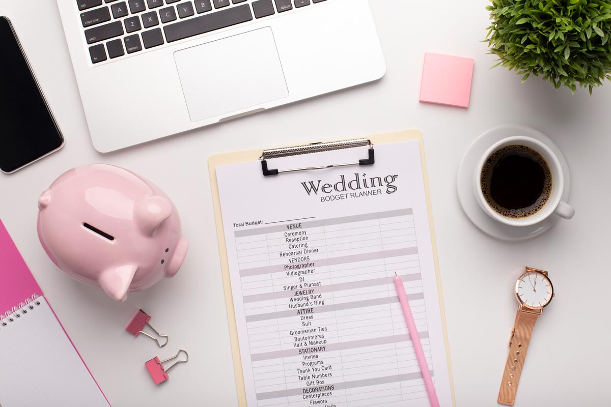 Wedding budget planner papers on white table with watch, full coffee cup, laptop, plant, and clips