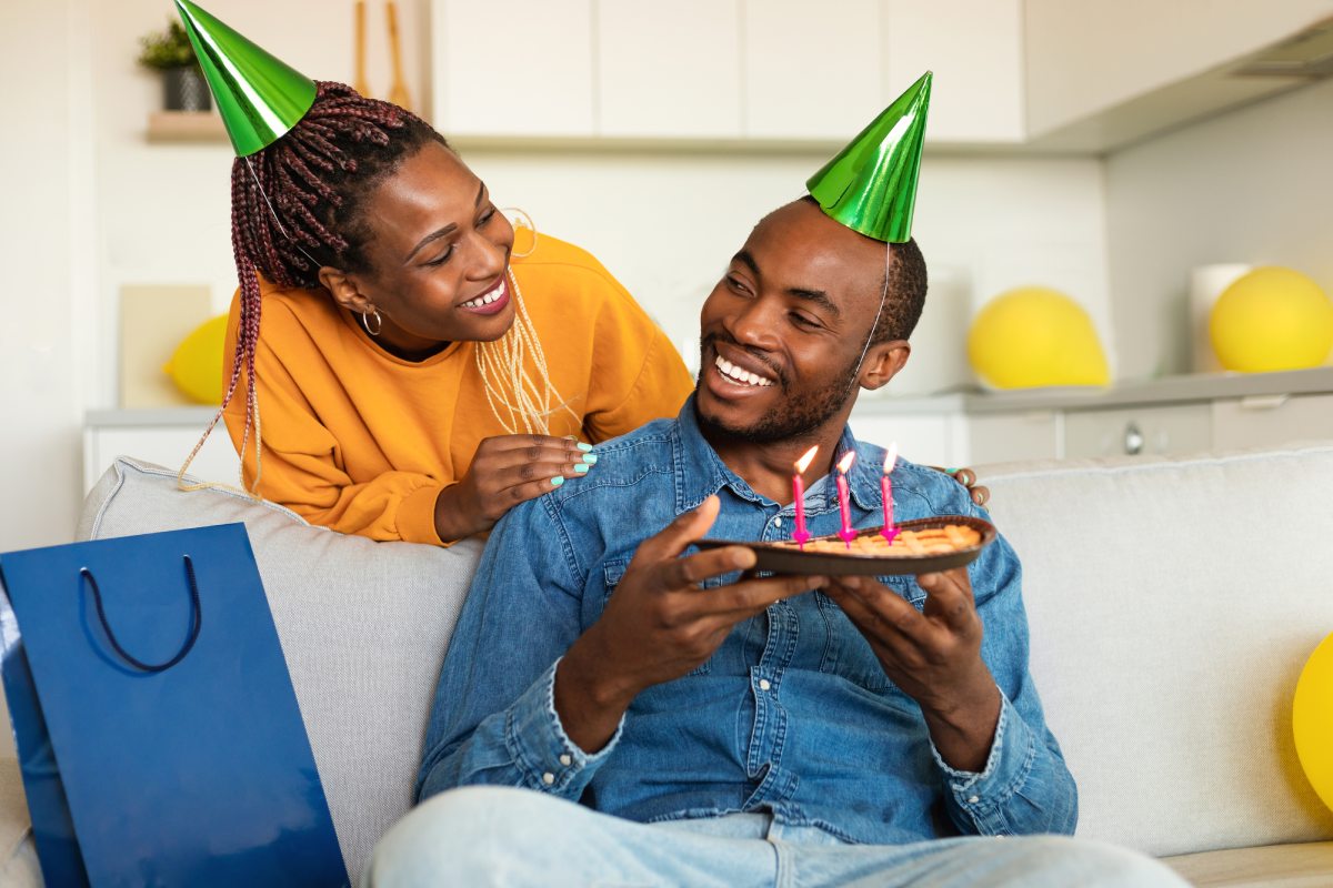 30-something black woman in matching green party hat with 30-something husband on the couch giving him waffles with candles on them for his birthday with a gift next to him