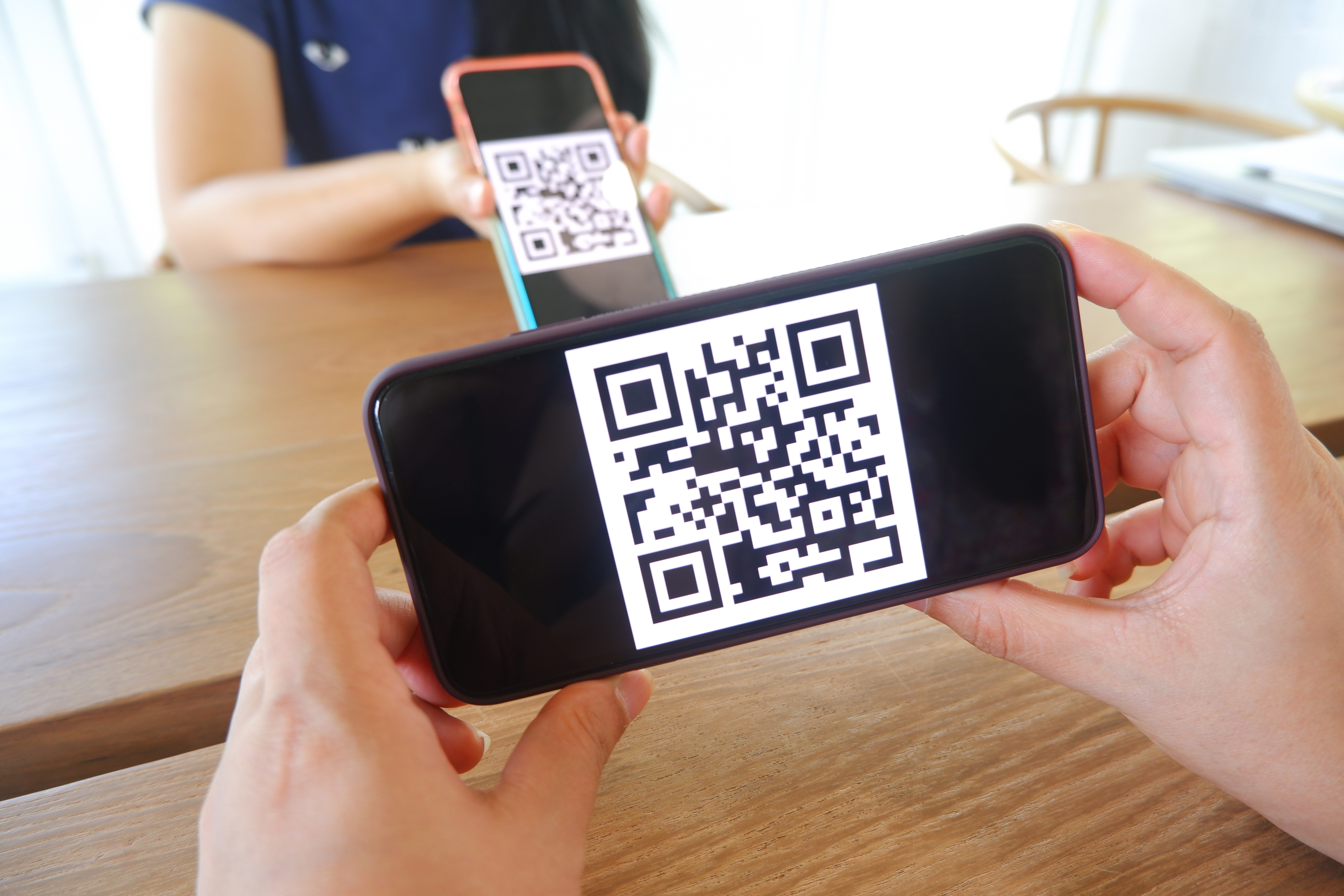  two sets of white hands hold cell phones with a QR codes as they sit on opposite sides of a table 