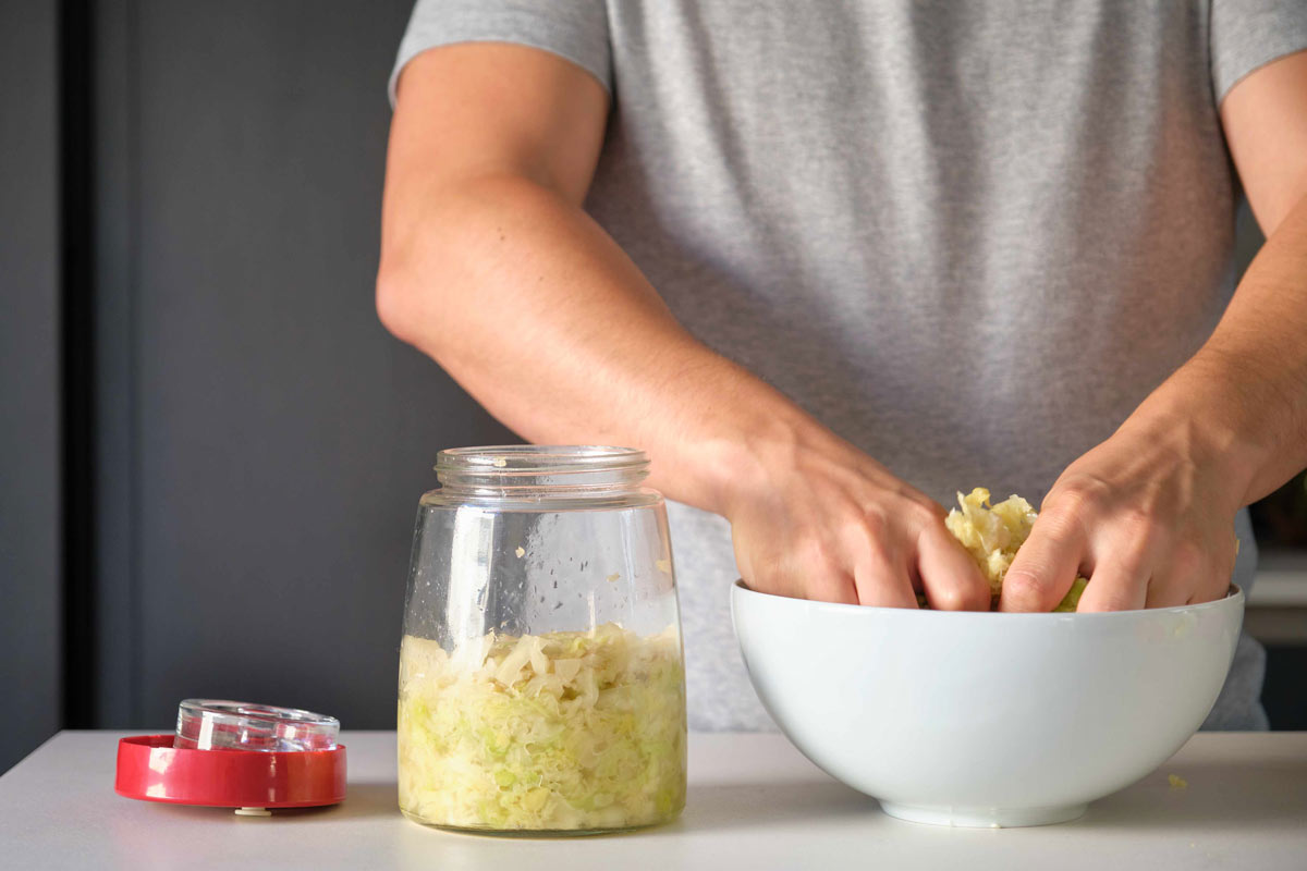  Man in a gray t-shirt preparing sauerkraut in a glass jar with his hands, showing a method of preserving food through fermenting.