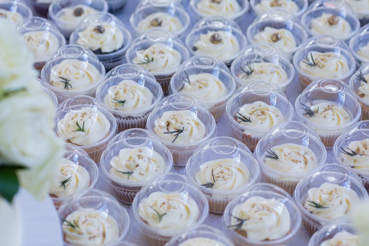 Many cupcakes with white frosting and green garnish in small to go containers 