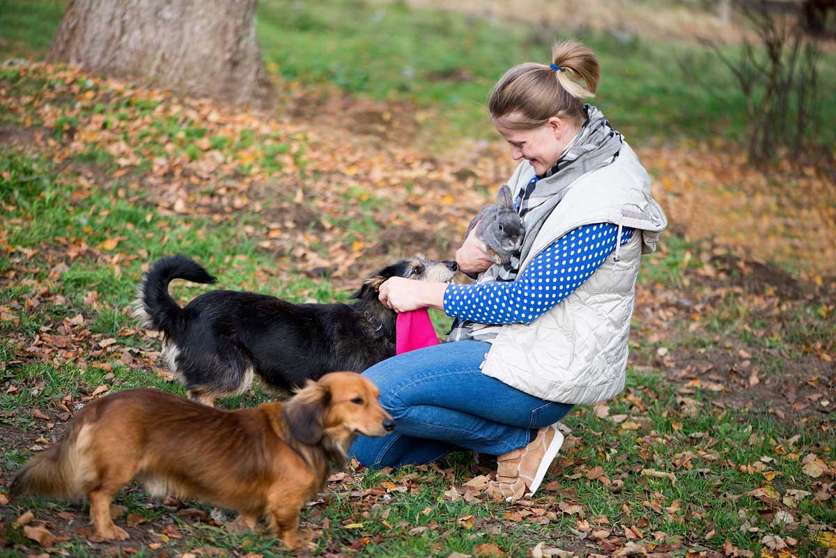 Woman kneeling down petting two dogs, while holding a bunny in the woods