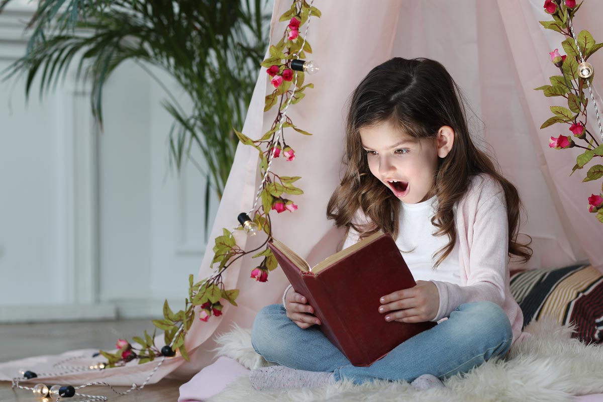 A white little girl sitting under a bed canopy reading a book with a shocked expressio