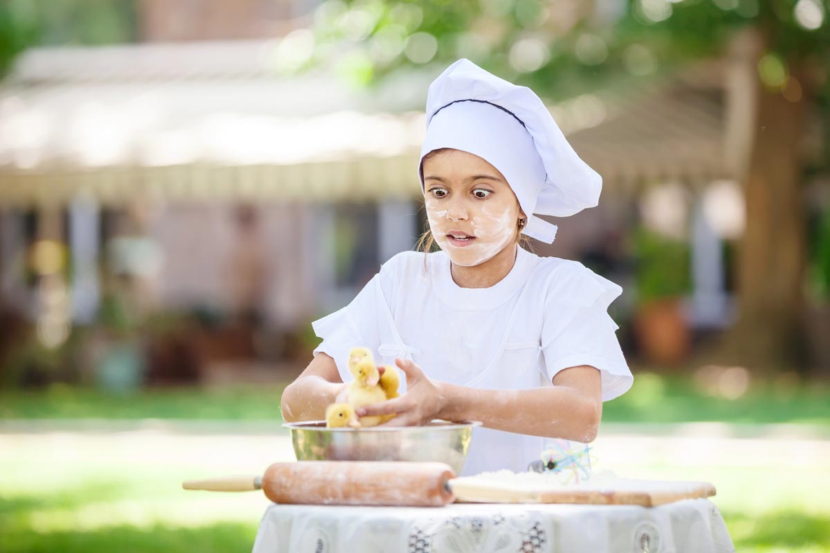 A little girl in a chef outfit with flour on her face mixing dough with her hands looking shocked