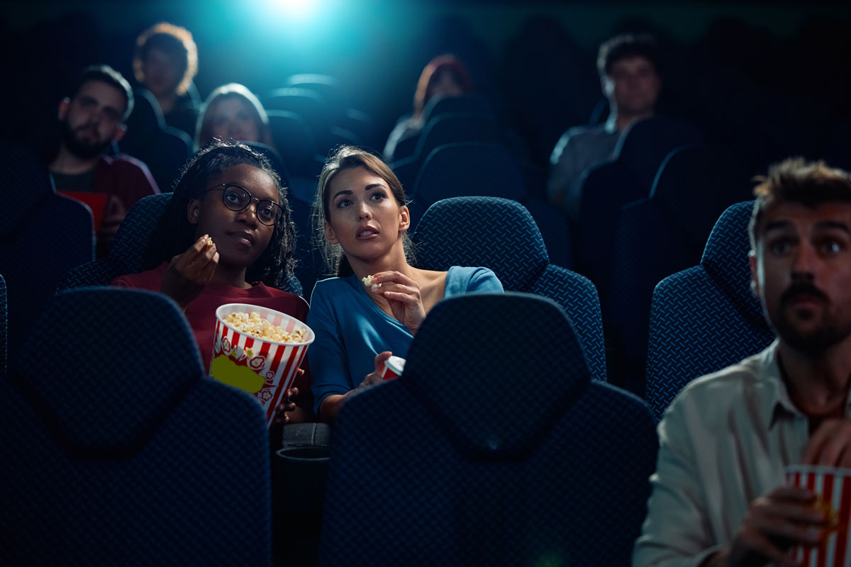 two women leaning in to each other while watching a movie in a movie theater with other people. 