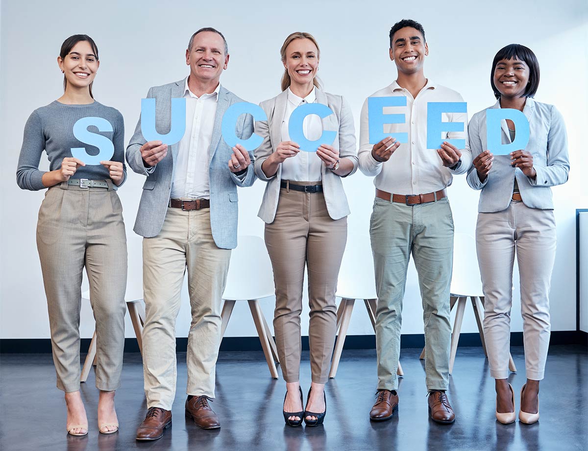 Group of multiracial adult professionals in a bare indoor office room smiling and holding up blue letters to spell the word “succeed”