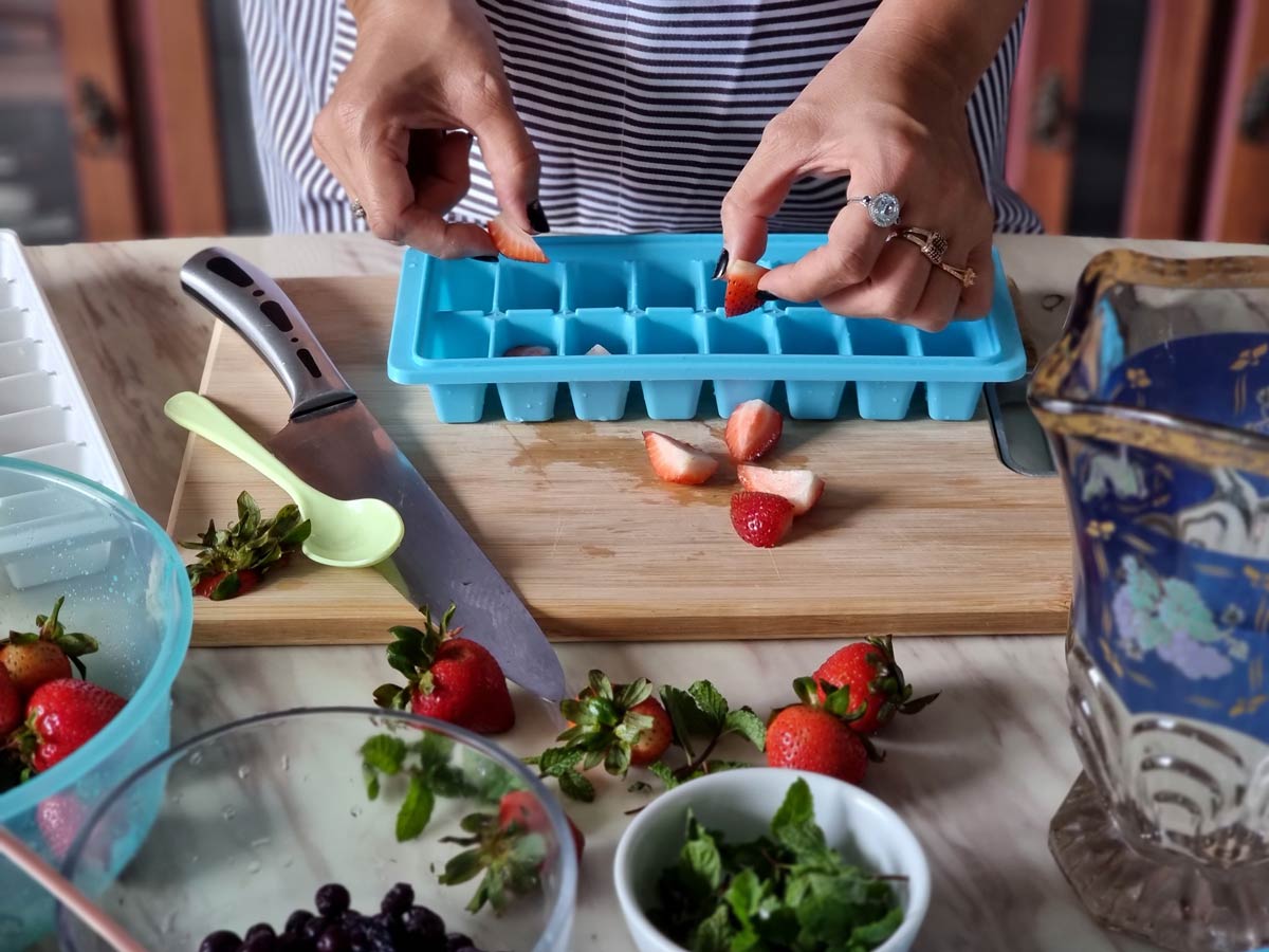 Close-up of white female hands placing cut-up strawberries in a blue ice tray that sits atop a wooden board surrounded by bowls of fruit, with a large cutting knife nearby. 