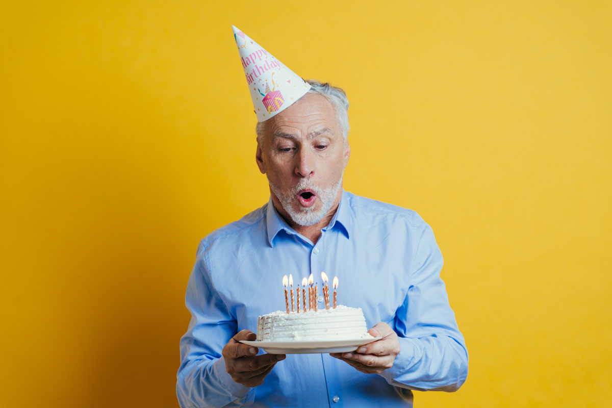 Older white man blowing out candles on his birthday cake, the man is wearing a birthday hat and the background is a yellow wall.