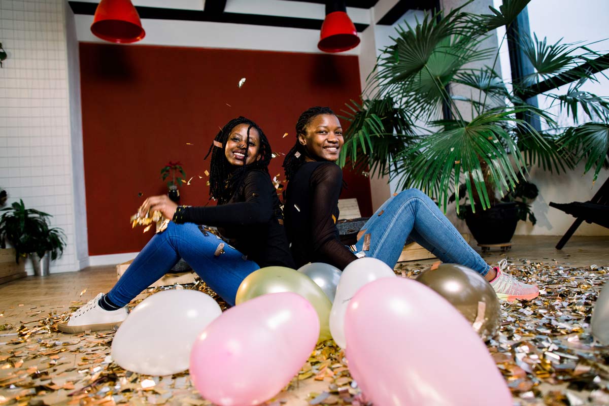 Two African American girls sitting on the floor with confetti and balloons smiling and happy