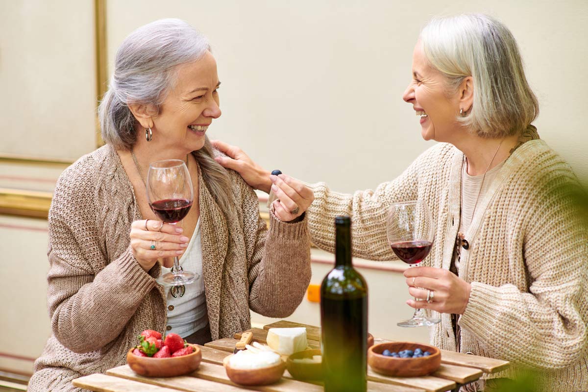 Two elderly women laughing in sweaters drinking wine and eating cheese and fruits