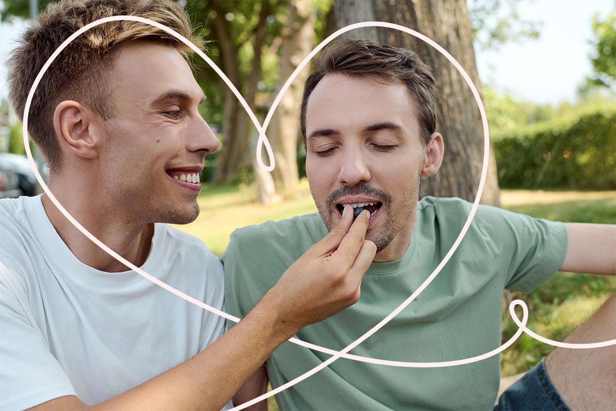  Two 20 something white men sit in a park with trees while one of them smiles and feeds the other.