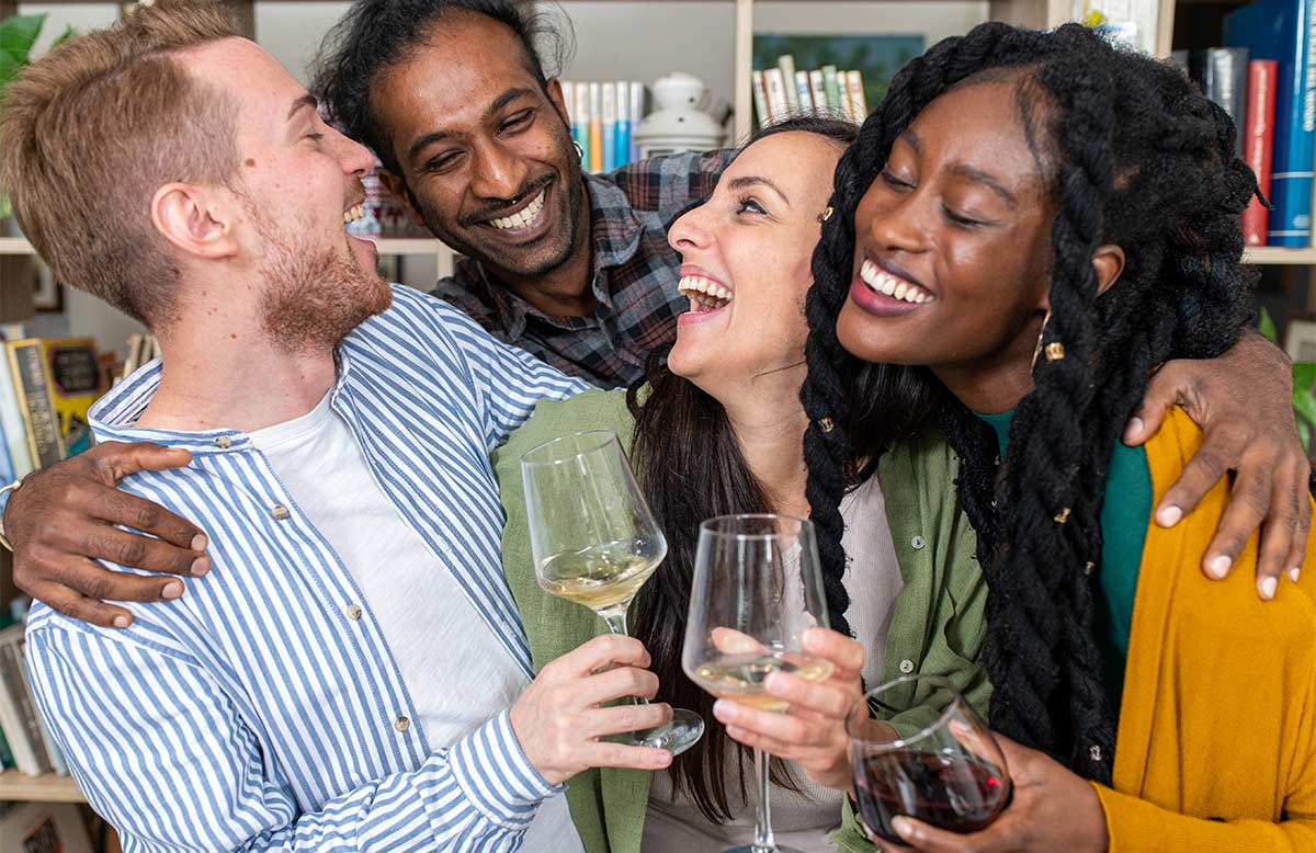 Multiracial group of adult friends in their 40s laugh and enjoy white and red wine while hugging indoors