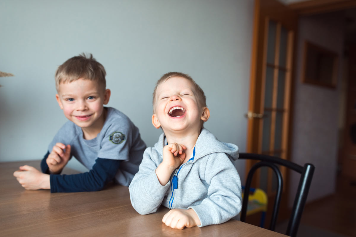  A young white boy sitting at a table throwing his head back and laughing with his older brother sitting behind him and smiling at the camera.