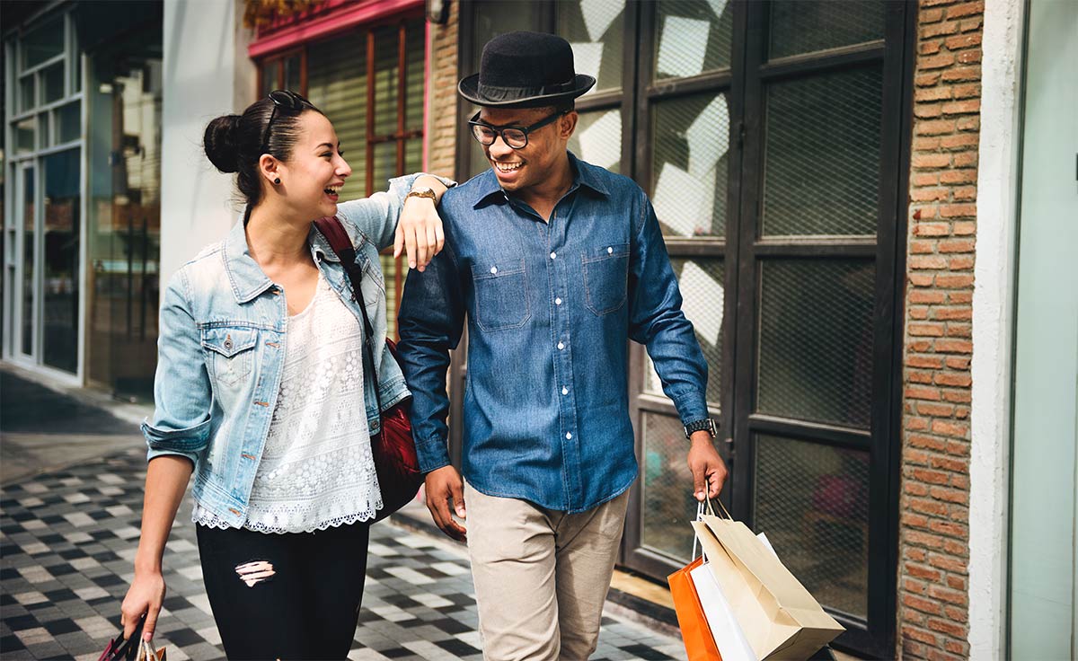 A young adult couple, where the man is black and the woman is white, are walking in the street, while the man is holding shopping bags.