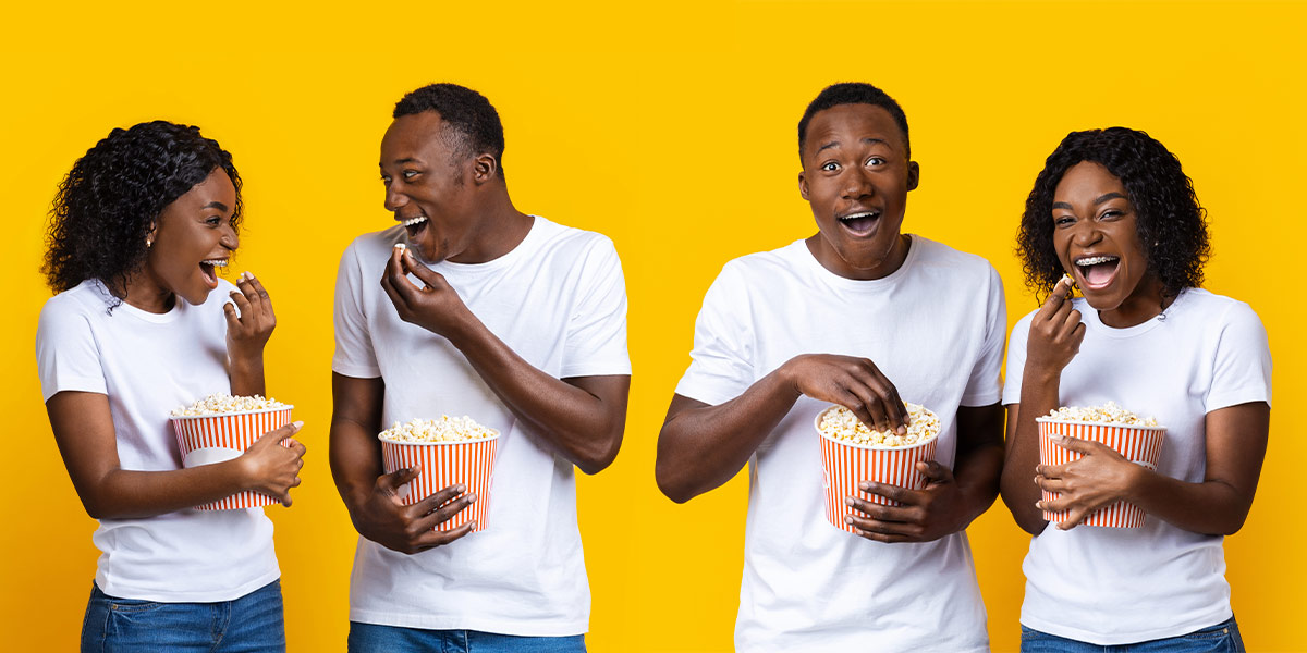 : A 20-something year old black couple are smiling and posing while eating popcorn, standing against a yellow wall.