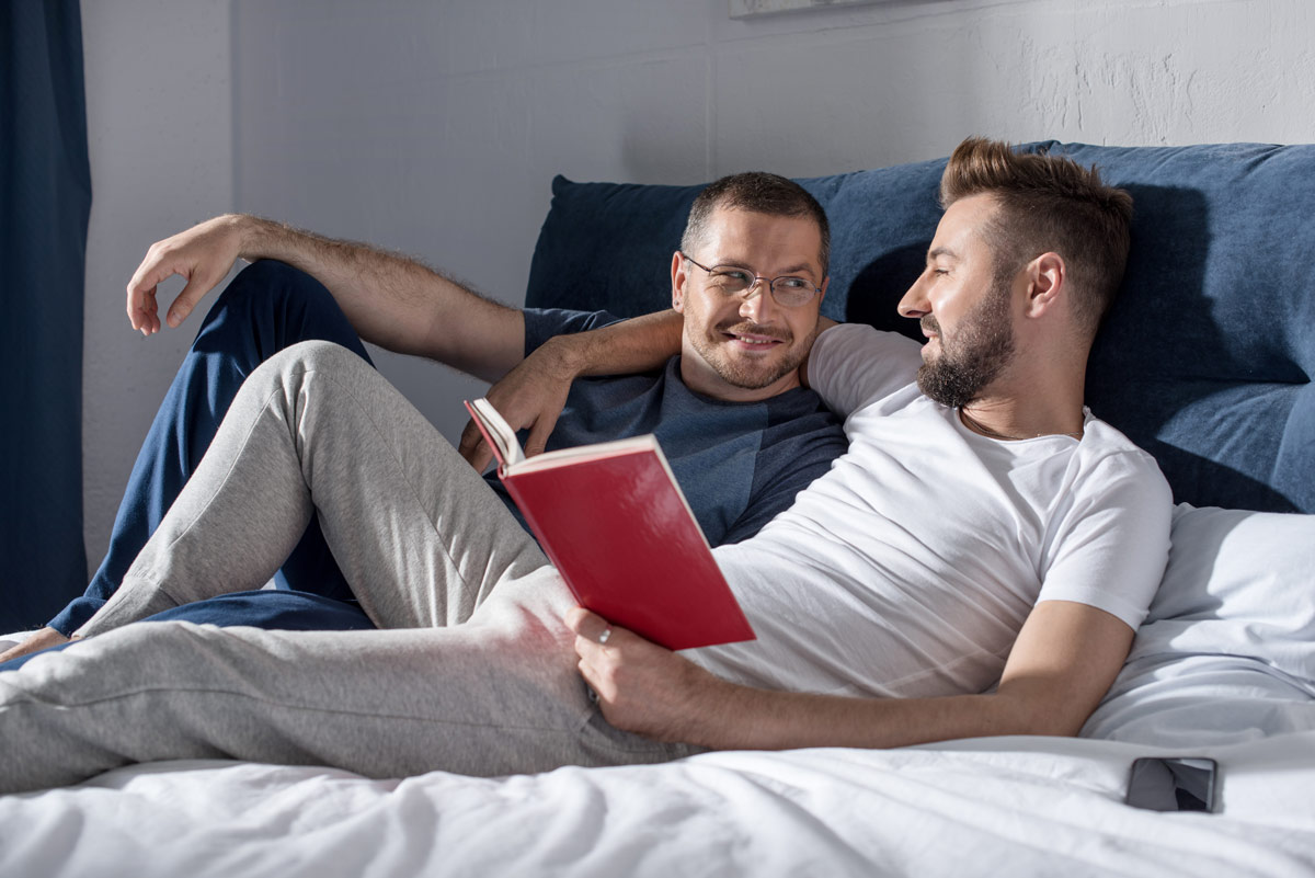 Two adult men, one White with a beard and the other white man with glasses, are relaxing together on a bed, smiling warmly at each other while one holds a book.