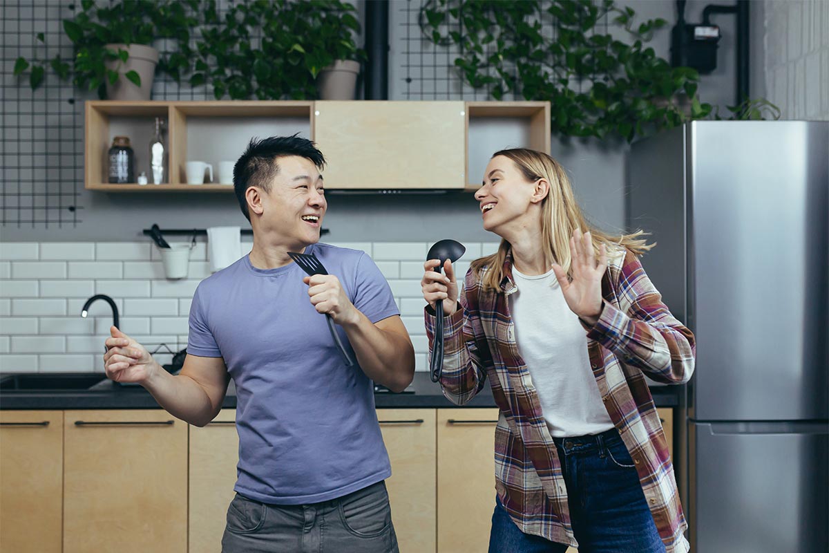 A young adult couple, the man is Asian and the woman is white, are singing in their kitchen, using soup ladles as a microphone. 