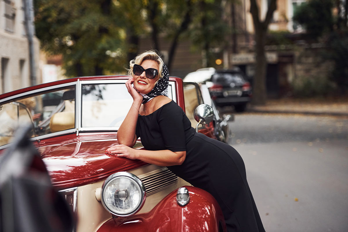 A smiling 60-year-old white woman wearing a black dress and a polka dot scarf with black sunglasses leans on a vintage car.